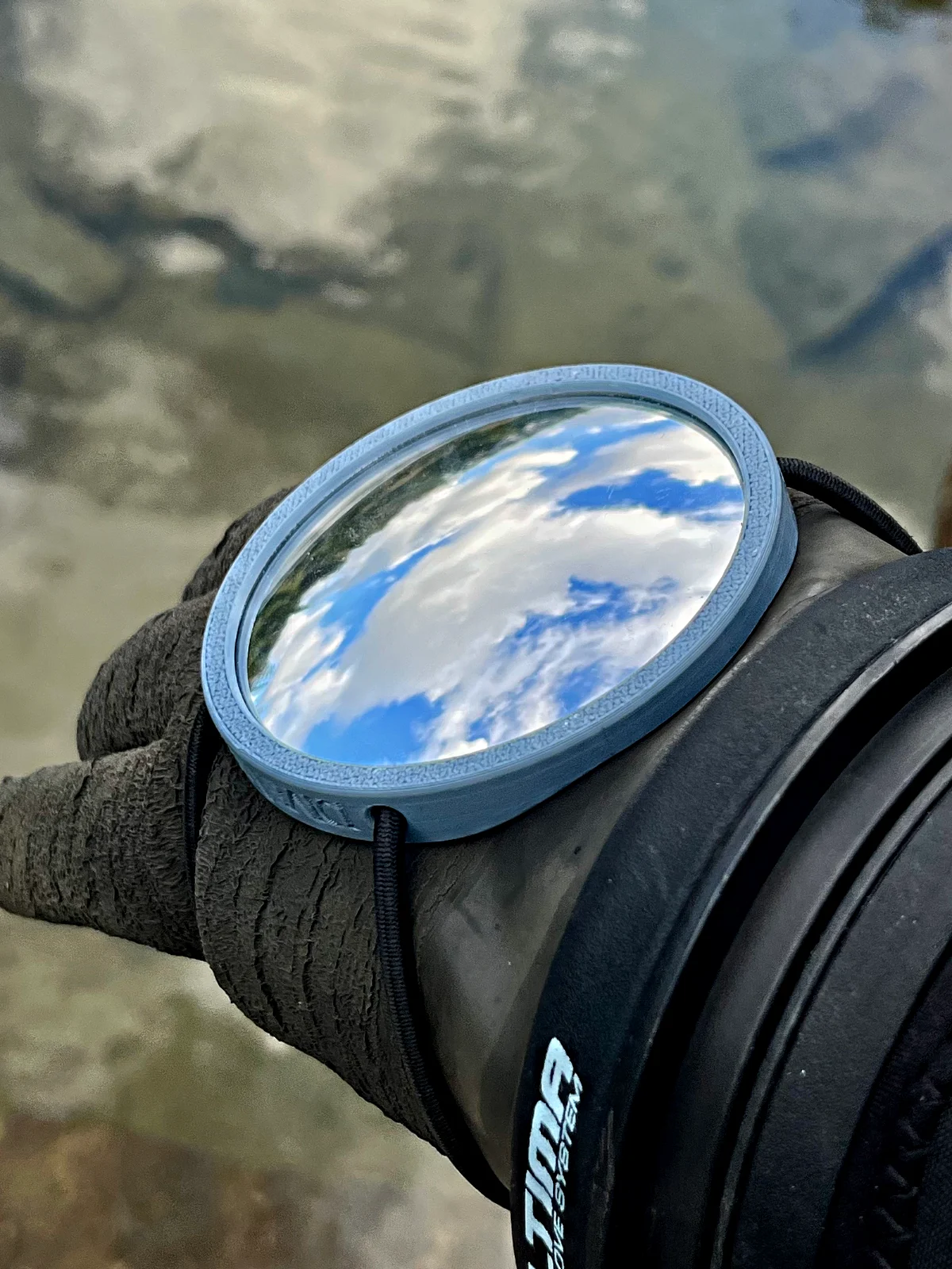 The Mirror 75 on a drysuit glove, clouds reflected, quarry water behind